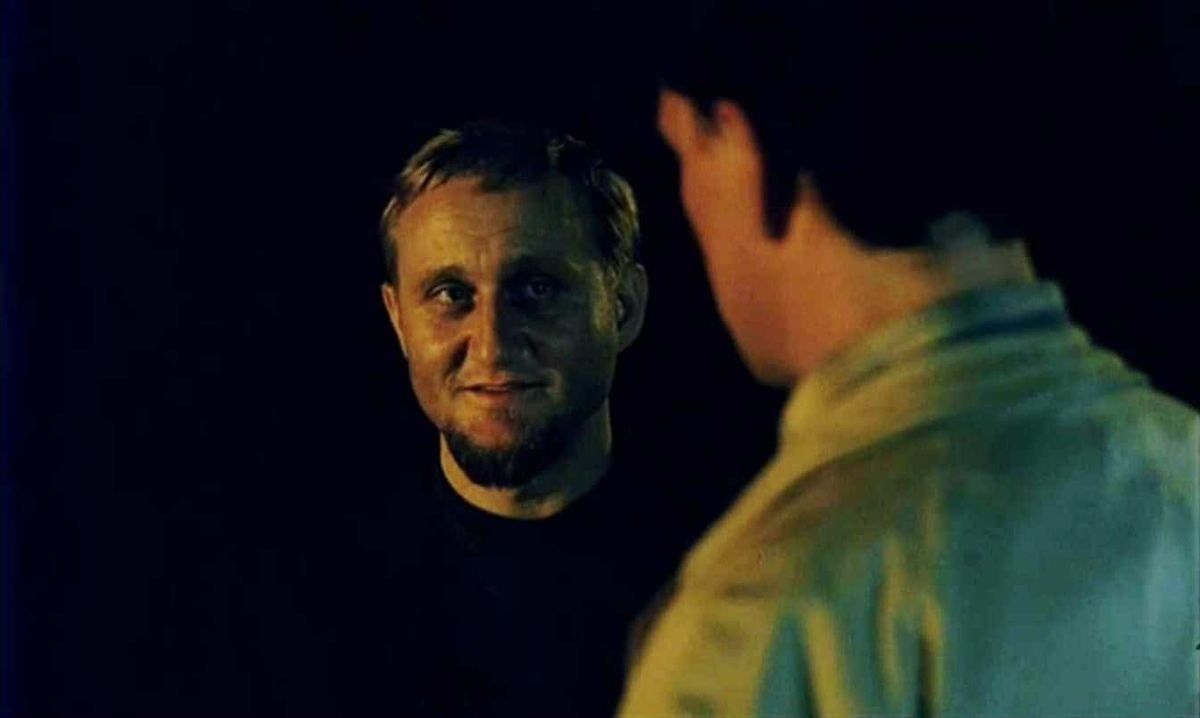 An evil man with a small beard smiles at another man against a black background in The Vanishing.